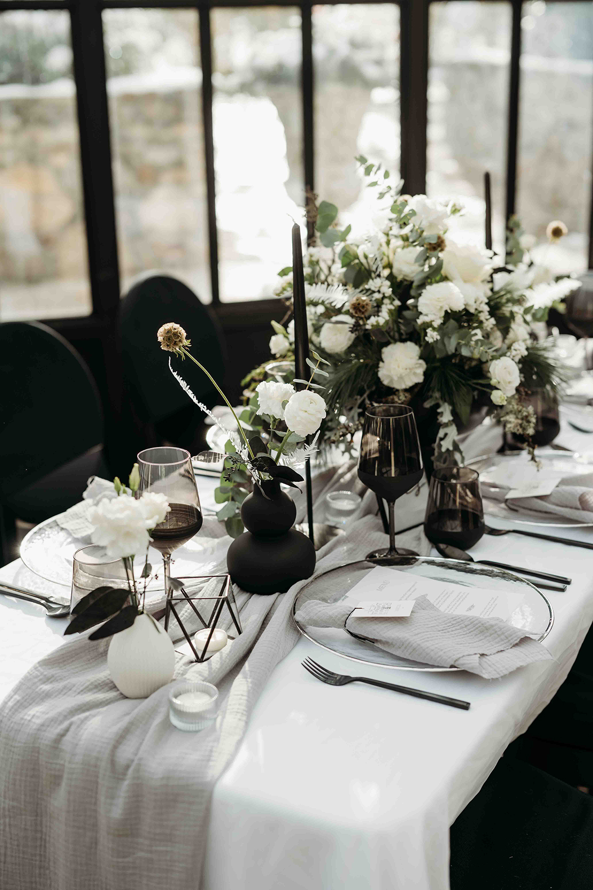 weiße und schwarze Hochzeitstafel, Tisch ist schön gedcket mit schwarzen Gläsern und Besteck, Teller sind in Glasoptik gehalten, ein großer Strauß mit weißen Rosen ziert die Mitte des Tisches