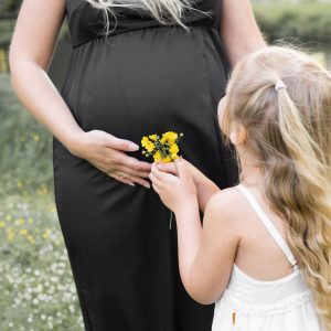 Mama in Vorfreude auf ihr Baby, Sie haltet ihren Bauch sie steht mit ihrer älteren Tochter die ihr gelbe Blumen reicht in frühlingshafter Landschaft