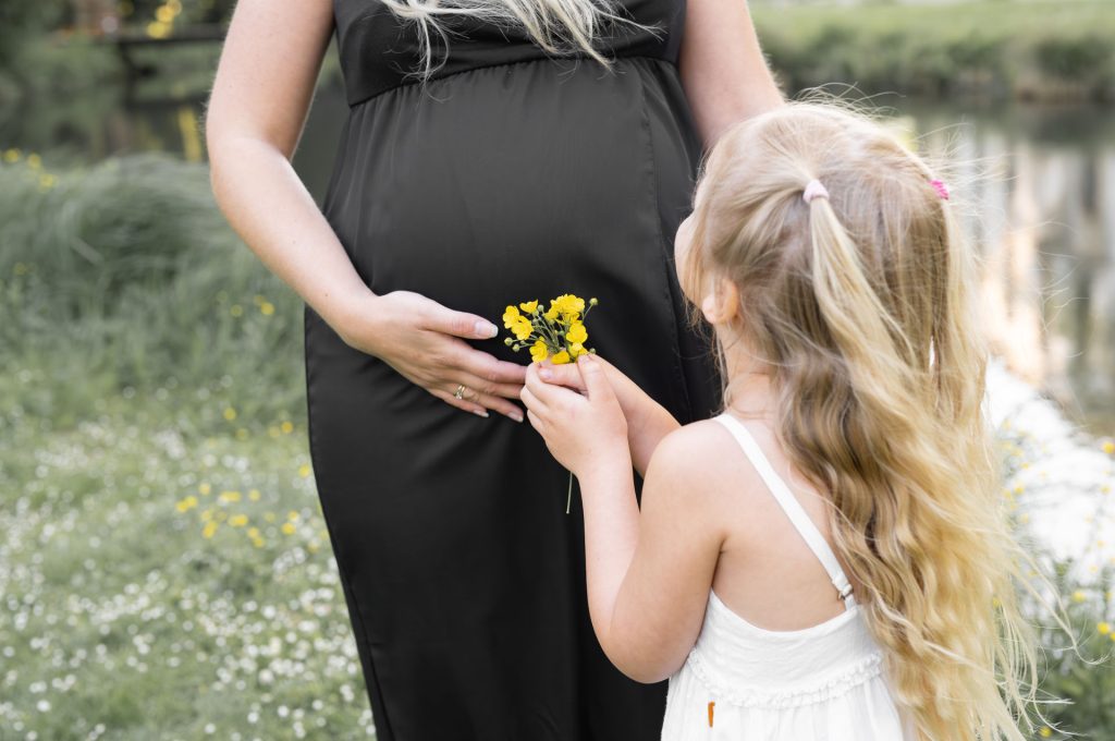 Mama in Vorfreude auf ihr Baby, Sie haltet ihren Bauch sie steht mit ihrer älteren Tochter die ihr gelbe Blumen reicht in frühlingshafter Landschaft